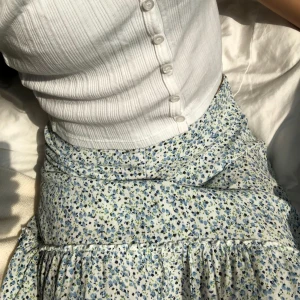 Blommig kjol med volang från Brandy Melville - Supersöt vit kjol med blått blommönster från Brandy Melville. Kjolen har en volang längst ner och dragkedja i sidan. Perfekt för en fräsch och somrig look🩷