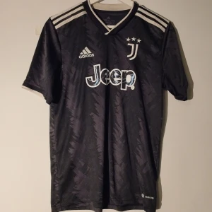 Juventus svart fotbollströja från Adidas - Juventus svart fotbollströja med vita detaljer och tre stjärnor ovanför klubbmärket. Adidas-logga och Jeep som sponsor på bröstet. Tröjan har korta ärmar och ett subtilt mönster i tyget. Perfekt för fans av Juventus. Storlek S. Obs inget namn eller nummer på ryggen. Kolla profilen för mer fotbollströjor och grisch kläder bland annat.