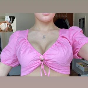 Rosa croppad topp med puffärmar och knyt - Säljer en söt rosa croppad topp med puffärmar och knyt framtill. Toppen har djup v-ringning och öppen rygg med knytband. Perfekt för varma dagar och ger en trendig look. Använd 0 ggr prislapp kvar, strl M men passar mig som har S för man kan knyta hur tajt man vill i ryggen 