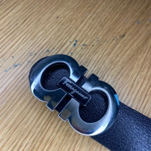 Svart skärp från Ferragamo med silverfärgat spänne - Säljer ett svart bälte från Ferragamo med ett stort, blankt silverfärgat spänne i metall. Bältet är gjort i skinn och har en klassisk, stilren design med Ferragamo-loggan på spännet.