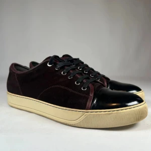 Svarta och vinröda sneakers med mocka och lackdetaljer - Snygga sneakers i vinröd mocka med svarta lackade tår och svarta snören. Skorna har en vit platt sula och rund tå. Storlek 44