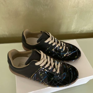 Maison Margiela sneakers - Unika svarta sneakers från Maison Margiela med färgstänk i blått, gult och vitt. Skorna har beige snörning, rund tå och platt sula. Perfekt för dig som vill sticka ut med en konstnärlig touch på fötterna. SIZE 41 MEN PASSAR 42 kommer med kvitto