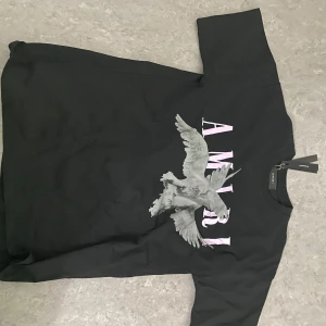 Amiri t shirt  - Säljer en svart t-shirt från Amiri med stort tryck av en fågel och text på bröstet. T-shirten har rund hals och korta ärmar. Perfekt för dig som gillar streetwear och vill ha något med cool design.