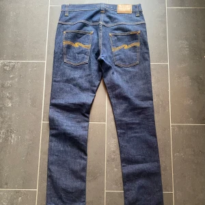 Mörkblå jeans från Nudie Jeans - Säljer ett par klassiska mörkblå jeans från Nudie Jeans med raka ben och snygga kontrastsömmar. Byxorna har fem fickor och Nudie-logga på bakfickan. Perfekta för dig som gillar stilrena jeans med bra passform.