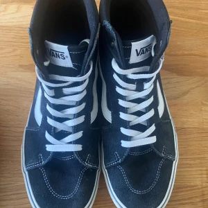 Svarta Vans high  - Säljer ett par klassiska svarta Vans high sneakers med beige sula och vit sidestripe. Skorna är tillverkade i mocka och syntet. Perfekta för dig som gillar streetstyle och vill ha en tidlös look.