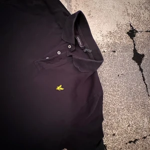Svart pikétröja från Lyle & Scott - Klassisk svart pikétröja från Lyle & Scott med gul broderad logga på bröstet. Tröjan har krage, knappar framtill och är tillverkad i bomull. Perfekt för en stilren och avslappnad look. Storlek 146-152