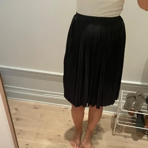 Svart plisserad kjol - Säljer en svart plisserad kjol med midjeresår. Kjolen har en klassisk A-linjeform och är knälång, vilket ger en snygg och tidlös look. Perfekt att styla med en enkel topp eller stickad tröja. Aldrig använd pga är ingen kjol-tjej
