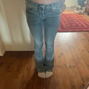 Blå jeansbyxor från True Religion - Säljer ett par klassiska blå jeans från True Religion med snygga detaljerade bakfickor och vita sömmar. Byxorna har låg midja och bootcut-stil, samt knappar och dragkedja framtill. Perfekta för dig som gillar en tidlös jeanslook.