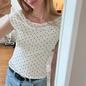 Vit prickig topp med draperad hals - Säljer en söt vit topp med svarta prickar och draperad halsringning. Toppen har korta ärmar och är figurnära, perfekt till jeans eller kjol. Enkel och stilren design som passar till många tillfällen. Köpt för 2 vecjor sedan. Använd 1 gång. Storlek Xs. 🩷