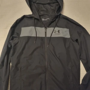 Svart vindjacka med huva från Under Armour - Svart vindjacka från Under Armour med grått bröstparti och logga. Jackan har huva med dragsko.