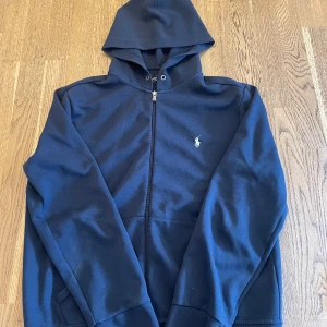 Mörkblå hoodie från Polo Ralph Lauren - Mörkblå hoodie med dragkedja från Polo Ralph Lauren. Klassisk design med huva och fickor framtill. Liten broderad logga i ljusblått på bröstet. Perfekt för en avslappnad stil.
