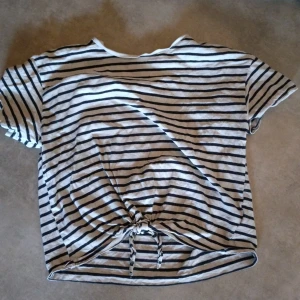 Randig t-shirt med knyt från H&M - Säljer en vit t-shirt med svarta ränder från H&M. Toppen har korta ärmar och en knytdetalj framtill som ger en snygg twist. Perfekt till jeans eller kjol för en avslappnad stil. Storlek 158/164 men är liten i storlek.