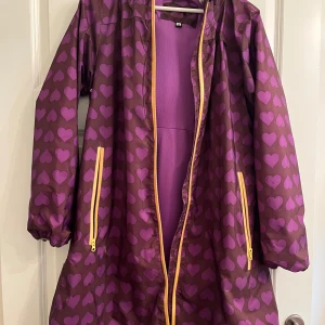 Lila regnjacka med hjärtmönster - Säljer en lila regnjacka med rosa hjärtan och gula dragkedjor. Jackan har huva, långa ärmar och två fickor framtill. Perfekt för regniga dagar och ger en färgglad touch till din outfit.