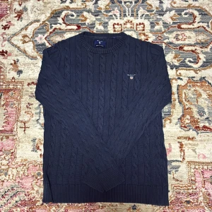 Mörkblå kabelstickad tröja från GANT - Säljer en klassisk mörkblå kabelstickad tröja från GANT med broderad logga på bröstet. Tröjan har rund halsringning och långa ärmar. Tillverkad i premium bomull för extra komfort.