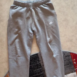 Grå mjukisbyxor från Nike - Säljer ett par klassiska grå mjukisbyxor från Nike med resår i midjan och liten vit Nike-logga på ena benet. Byxorna har en bakficka och är perfekta för chill eller träning. Superbekväma och stilrena!