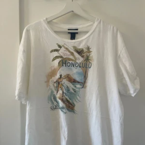 Vit t-shirt  Polo Ralph Lauren - Vit t-shirt från Polo Ralph Lauren med färgglatt tryck av surfare och palmer samt texten 'Trip to Honolulu' på bröstet. Rund halsringning och normal passform. Tillverkad i 100% bomull. För endast 159kr
