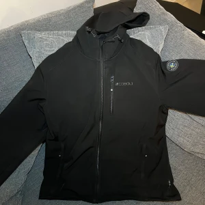 Svart softshelljacka med huva från Cobalt - Svart softshelljacka från Cobalt med huva och dragkedja framtill. Jackan har en bröstficka med dragkedja, två sidofickor och en patch på ärmen. Perfekt för kyliga dagar och enkel att matcha. Svensk flagga sidan av ärmen , Jackan har tappat lite av dina bokstäver vid CO i Cobolt*