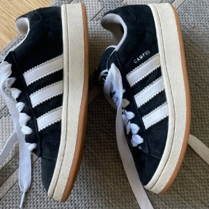 Svarta Adidas Campus sneakers med vita ränder - Säljer ett par klassiska Adidas Campus sneakers i svart mocka med vita ränder och vit sula. Skorna har breda vita skosnören och gummisula med brun undersida. Perfekta för en sportig och stilren look.