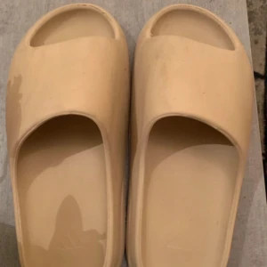 Beige Adidas Yeezy Slide sandaler - Säljer ett par beige Adidas Yeezy Slide sandaler. De har en minimalistisk design med öppen tå och platt sula, tillverkade i syntetmaterial. Perfekta för sommaren och riktigt sköna att glida runt i.