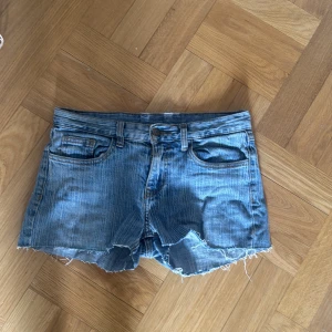 Blå jeansshorts med fransar - Säljer ett par klassiska blå jeansshorts med råa fransade kanter och fem fickor. Shortsen har bälteshällor och knappgylf. Perfekta för varma dagar och enkel att matcha med olika toppar.