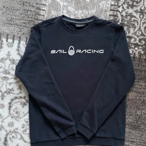 Svart sweatshirt från Sail Racing - Säljer en svart sweatshirt från Sail Racing med vit logotyp på bröstet. Tröjan har rund halsringning och långa ärmar. Perfekt för en stilren och sportig look.
