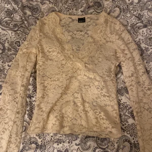 Beige spetsblus från Gina Tricot - Säljer en beige långärmad spetsblus från Gina Tricot i storlek XS. Blusen har v-ringning och är helt täckt av blommigt spetsmönster, vilket ger en romantisk och feminin look.