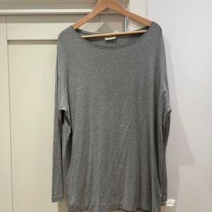 Grå långärmad tröja från By Malene Birger - En enkel och stilren grå långärmad tröja från By Malene Birger. Tröjan har en rund halsringning och är tillverkad i en mjuk viskosblandning som känns skön mot huden. Perfekt för en avslappnad och snygg look.