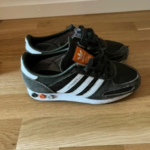 Adidas LA Trainer sneakers i svart mocka - Säljer ett par Adidas LA Trainer sneakers i svart mocka och textil med vita detaljer och klassiska tre ränder på sidan. Skorna har snörning och en vit sula med färgade pluggar. Perfekta för dig som gillar retrostil.