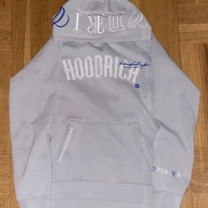 Grå hoodie från Hoodrich - Säljer en grå hoodie från Hoodrich med vit broderad logga och blå detaljer på bröstet och luvan. Tröjan har magficka och ribbade muddar vid ärmslut och nederkant. Perfekt för en avslappnad stil.
