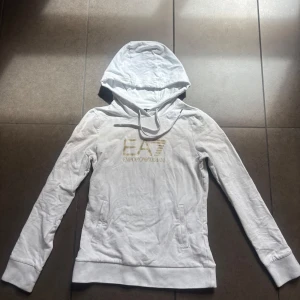 Vit hoodie från Emporio Armani EA7 - Säljer en vit hoodie från Emporio Armani EA7 med guldig logga framtill. Tröjan har huva, långa ärmar och ribbade muddar vid ärmslut och nederkant. Perfekt för en stilren och sportig look. Använd fåtal gånger. Nypris: 1 400kr