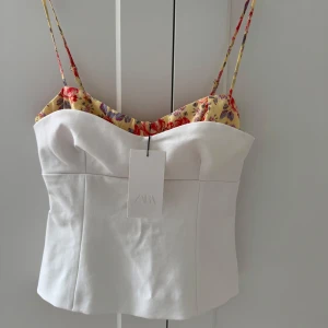 Vit korsettopp från ZARA - Vit korsettopp från Zara med blommiga detaljer. Oanvänd i storlek M, men är ganska liten i storleken så passar även S.