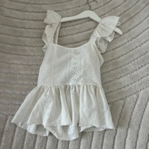 Vit volangtopp med broderade detaljer - Söt vit topp med volanger på axelbanden och nertill, samt broderade hålmönster. Toppen har knytning i ryggen och är ärmlös med en luftig och somrig känsla.