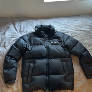 Svart dunjacka från Sail Racing - Säljer en svart dunjacka från Sail Racing med hög krage och dragkedja framtill. Jackan har två stora fickor på sidorna och logga på bröstet. Perfekt för kyliga dagar och riktigt skön att bära.