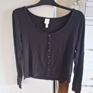 Svart ribbad tröja från H&M - Säljer en svart ribbad tröja från H&M i storlek XL. Koftan har långa ärmar, rundad urringning och knappar framtill. Perfekt att slänga över en topp eller klänning för en enkel och stilren look.