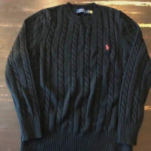 Svart kabelstickad tröja från Polo Ralph Lauren - Kabelstickad svart tröja från Polo Ralph Lauren med rund halsringning och rött broderat logotyp på bröstet. Tröjan har långa ärmar och ribbade muddar vid ärmslut och nederkant. Perfekt för en stilren look.