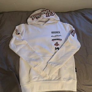 Vit hoodie från Hoodrich med broderade detaljer - Säljer en vit hoodie från Hoodrich med flera broderade loggor och tryck på bröstet och ärmarna. Stor broderad text på huvan. Hoodien har normal passform och är perfekt för dig som gillar streetwear.