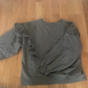 Grön sweatshirt med volang från Gina Tricot/ stl 154-164 - Säljer en grå sweatshirt från Gina Tricot med volangdetaljer längs axlarna och ärmarna. Tröjan har rund halsringning och långa ärmar med muddar. Perfekt för en avslappnad och trendig stil.