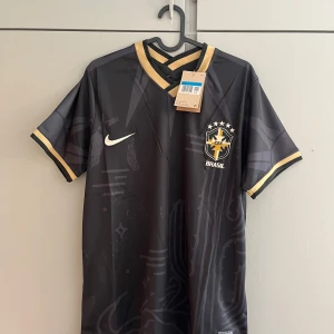 Svart Brasilien fotbollströja med guldiga detaljer - Snygg svart Brasilien fotbollströja från Nike med guldiga detaljer och emblem på bröstet. Tröjan har korta ärmar, V-ringad krage med guldkant och diskreta mönster i tyget. Perfekt för dig som vill sticka ut på planen eller på läktaren.