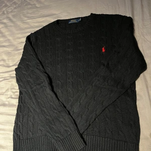 Svart kabelstickad tröja från Polo Ralph Lauren - Kabelstickad svart tröja från Polo Ralph Lauren med rund halsringning och röd broderad logga på bröstet. Klassisk och stilren design, perfekt för lager-på-lager. Mjuk och bekväm med ribbade muddar, storlek L men passar även M. Öppen för prisförslag!
