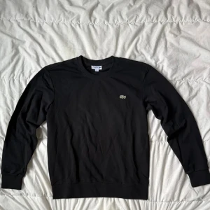 Svart sweatshirt från Lacoste - Klassisk svart sweatshirt från Lacoste med rund halsringning och det ikoniska krokodilmärket broderat på bröstet. Tröjan har långa ärmar och är tillverkad i mjuk bomull för en bekväm känsla.