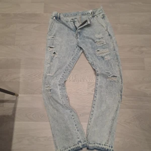 Ljusblå slitna jeans från Sumwon - Säljer ett par ljusblå jeans från Sumwon med flera slitningar och hål på framsidan. Jeansen har klassisk femficksdesign och normal passform. Perfekta för en avslappnad och trendig look.