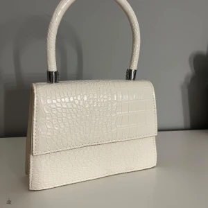 Vit handväska med krokodilpräglat mönster - Snygg vit handväska med krokodilpräglat mönster och stilrent handtag. Väskan har en klassisk form och är perfekt för att lyfta vilken outfit som helst. Enkel att matcha och ger en lyxig känsla.