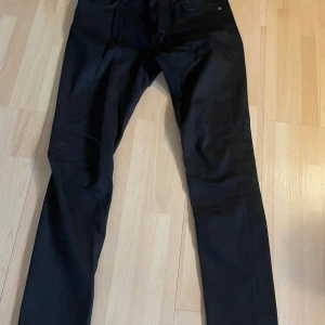 Svarta jeans från Nudie Jeans - Säljer ett par svarta jeans från Nudie Jeans i klassisk rak modell. Jeansen har fem fickor, bälteshällor och stängs med dragkedja och knapp. Perfekta för dig som gillar stilrena och tidlösa byxor.