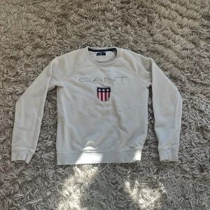 Vit sweatshirt från GANT - Vit sweatshirt från GANT med broderad logotyp och sköld på bröstet. Tröjan har rund halsringning och långa ärmar. Klassisk och stilren design med ribbade muddar vid ärmslut och nederkant.