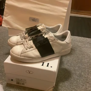 Valentino Open sneakers med svart detalj - Säljer ett par vita Valentino Open sneakers med bred svart läderdetalj över mitten. Klassisk låg modell med rund tå och platt sula. Skorna har diskret logga på sidan och vita snören. Kommer med originalkartong och dustbag. Kom med bud!! Byten kan vara intressant. Äkthetsbevis finns. 