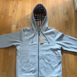 Grå zip hoodie från Burberry - Säljer en grå zip hoodie från Burberry med klassiskt rutigt foder i huvan. Jackan har dragkedja framtill, två fickor och justerbar huva. Perfekt för dig som gillar stilren design med en touch av lyx.
