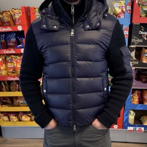 Mörkblå dunväst med stickade ärmar från Moncler - Snygg mörkblå dunväst från Moncler med stickade ärmar och huva. Västen har dragkedja framtill, två fickor med knappar och en ficka med Moncler-logga på ärmen. Perfekt för lager-på-lager och stilren look.