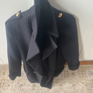 Marinblå skepparkappa detaljer  - Säljer en marinblå kappa från Zara med bred krage och dekorativa guldknappar på axlar och framtill. Jackan har lång ärm och två fickor framtill. Perfekt för kyligare dagar och ger en stilren look. Passar medium o large 