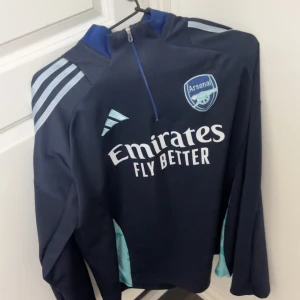 Arsenal träningströja med halv dragkedja - Mörkblå Arsenal träningströja från Adidas med halv dragkedja och ljusblå detaljer. Tryckt klubbmärke och sponsorlogga på bröstet samt klassiska Adidas-ränder på axlarna. Perfekt för fotbollsträning eller som supporterplagg.