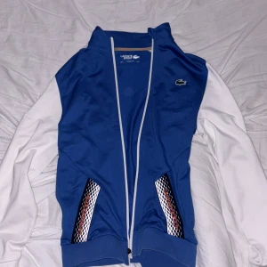 Blå och vit sportig jacka från Lacoste - Säljer en blå och vit sportjacka från Lacoste med dragkedja och klassisk krokodillogga på bröstet. Jackan har långa ärmar, vita detaljer och mönstrade fickor med racing-inspirerat tryck. Perfekt för en sportig look.
